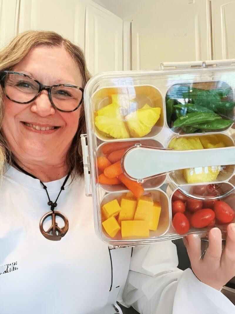Person holding a bento box with compartments of fruits and vegetables in a kitchen setting.